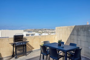 A rooftop terrace with a barbecue grill, a table with chairs, and a view of the cityscape. - Gozo Sannat Penthouse 3BDR (Sannat)