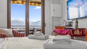 Quarto de hotel com uma cama, uma mesa de jantar com toalha vermelha, uma televisão exibindo uma vista de montanhas e vista para montanhas pela janela.