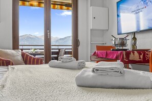 Ein Hotelzimmer mit einem Bett, einem Esstisch mit roter Tischdecke, einem Fernseher, der eine Bergblick zeigt, und Blick auf Berge durch das Fenster.