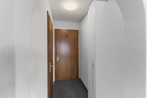 Ein schmaler Flur mit einer Holztür, weißen Wänden mit Struktur und einer Deckenleuchte.