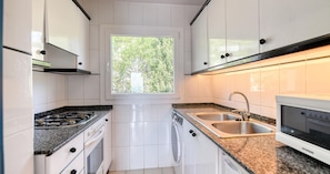 A modern kitchen with white cabinets, a gas stove, a built-in oven, a double sink, and a window providing natural light. - Sea-view apartment in El Golfet (Calella de Palafrugell)
