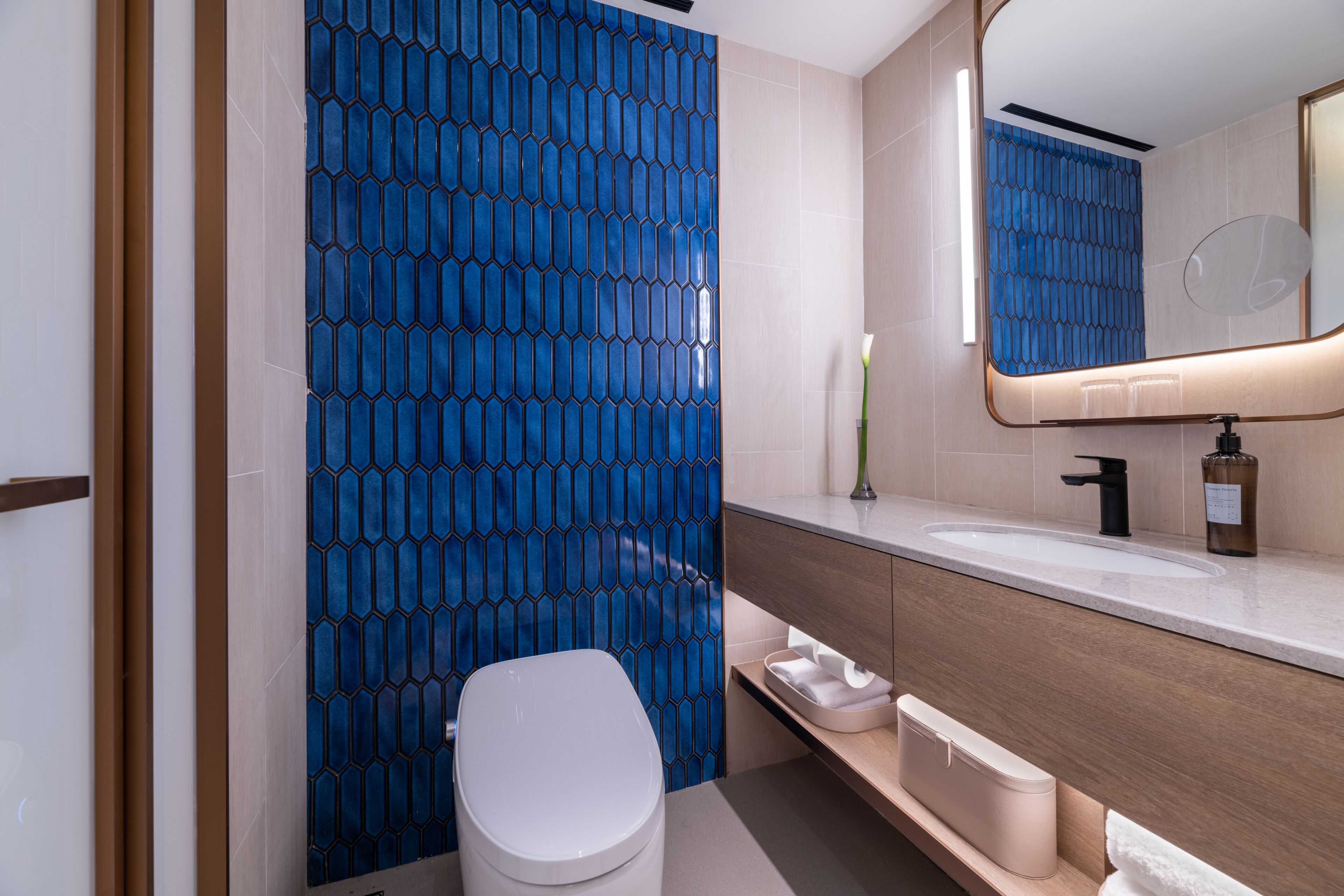 A modern bathroom with a blue hexagonal tile wall, a white toilet, a vanity with a mirror, and a sink.