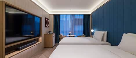A hotel room with a large flat-screen TV, a desk with a lamp, two beds, and a view of the outdoors through sheer curtains.