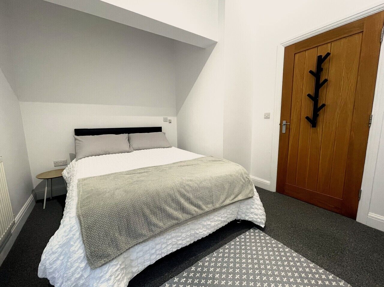 A bedroom with a bed, a bedside table, a chair, a door, and a patterned rug. - Walk to Everything | Sleeps 4 | 2 Bed | Bold St (Merseyside)