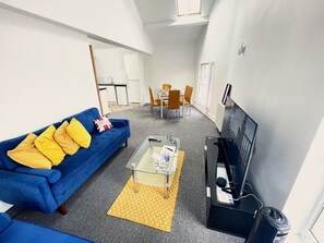 A living room with a blue sofa, a glass coffee table, a TV, and a dining area in the background. - Walk to Everything | Sleeps 4 | 2 Bed | Bold St (Merseyside)