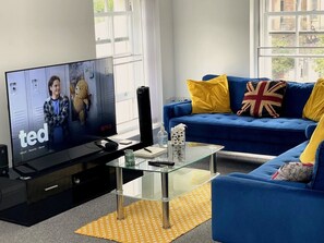 A modern living room with a flat-screen TV, a glass coffee table, a blue sofa with yellow and red cushions, and a window with a view. - Walk to Everything | Sleeps 4 | 2 Bed | Bold St (Merseyside)