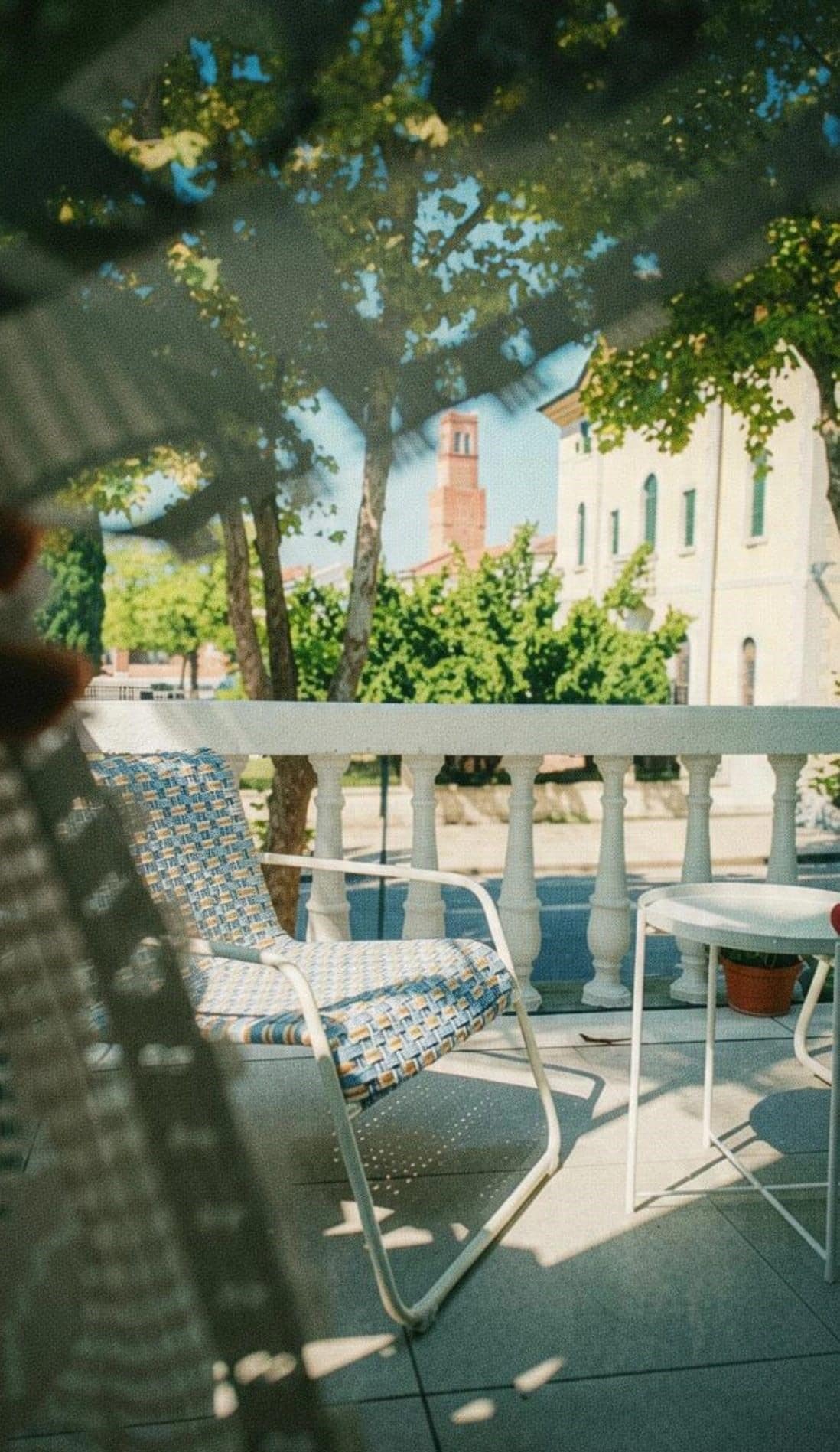 Eine Terrasse mit einem Metallstuhl und einem kleinen Tisch, einem weißen Geländer und einem Gebäude mit einem Turm im Hintergrund.
