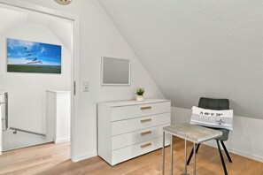 Ein modernes Zimmer mit einer weißen Kommode, einem kleinen Tisch, einem Stuhl, einem Gemälde und einem Spiegel an der Wand.