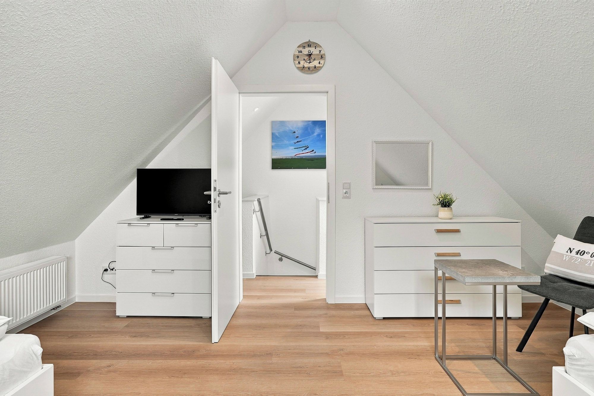 Ein modernes Schlafzimmer im Dachgeschoss, eingerichtet in Weiß, mit Fernseher, einem Gemälde, einem Spiegel und einem kleinen Tisch.