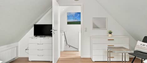 Ein modernes Schlafzimmer im Dachgeschoss, eingerichtet in Weiß, mit Fernseher, einem Gemälde, einem Spiegel und einem kleinen Tisch.