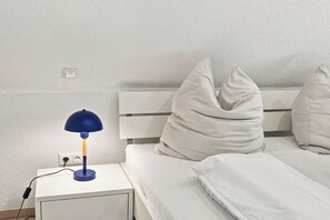 A bedroom with a white bed, a nightstand with a lamp, and a power outlet. - Lüttje Sünn - Your Oasis of Well-Being in Norddeich (Norddeich)