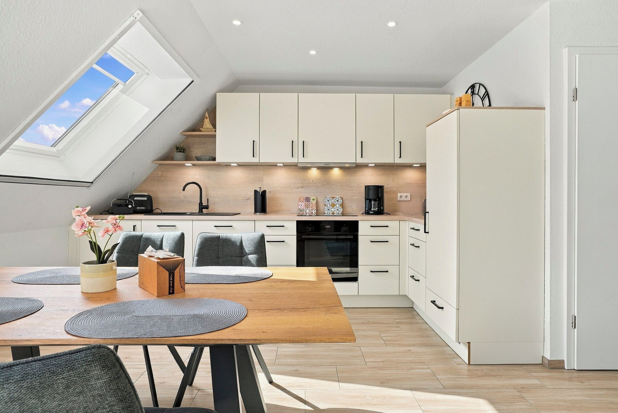 A modern kitchen with white cabinets, a wooden dining table, and a skylight.