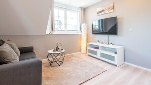 Ein modernes Wohnzimmer mit einem grauen Sofa, einer weißen TV-Konsole, einem kleinen runden Tisch mit einer Vase und einem großen Fenster mit Vorhängen.