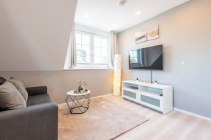 A modern living room with a grey sofa, a white TV stand, a small round table with a vase, and a large window with curtains. - Vacation Home "Westerlön Hüs 1999" (Sylt)