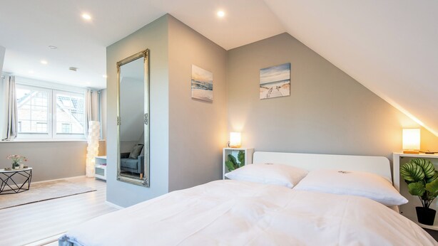 Ein Schlafzimmer mit einem Bett, einem Spiegel, zwei Nachttischlampen, einem Fenster und einer Pflanze.