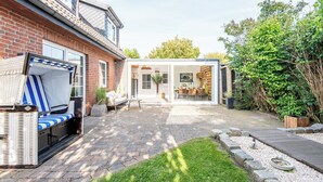 Eine Terrasse mit einer Rattan-Couch, einem durch eine Glaswand geschützten Essbereich und einem Ziegelhaus.