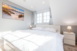 A bedroom with a large bed, a nightstand, a lamp, and a window with curtains. - Vacation Home "Westerlön Hüs 1999" (Sylt)