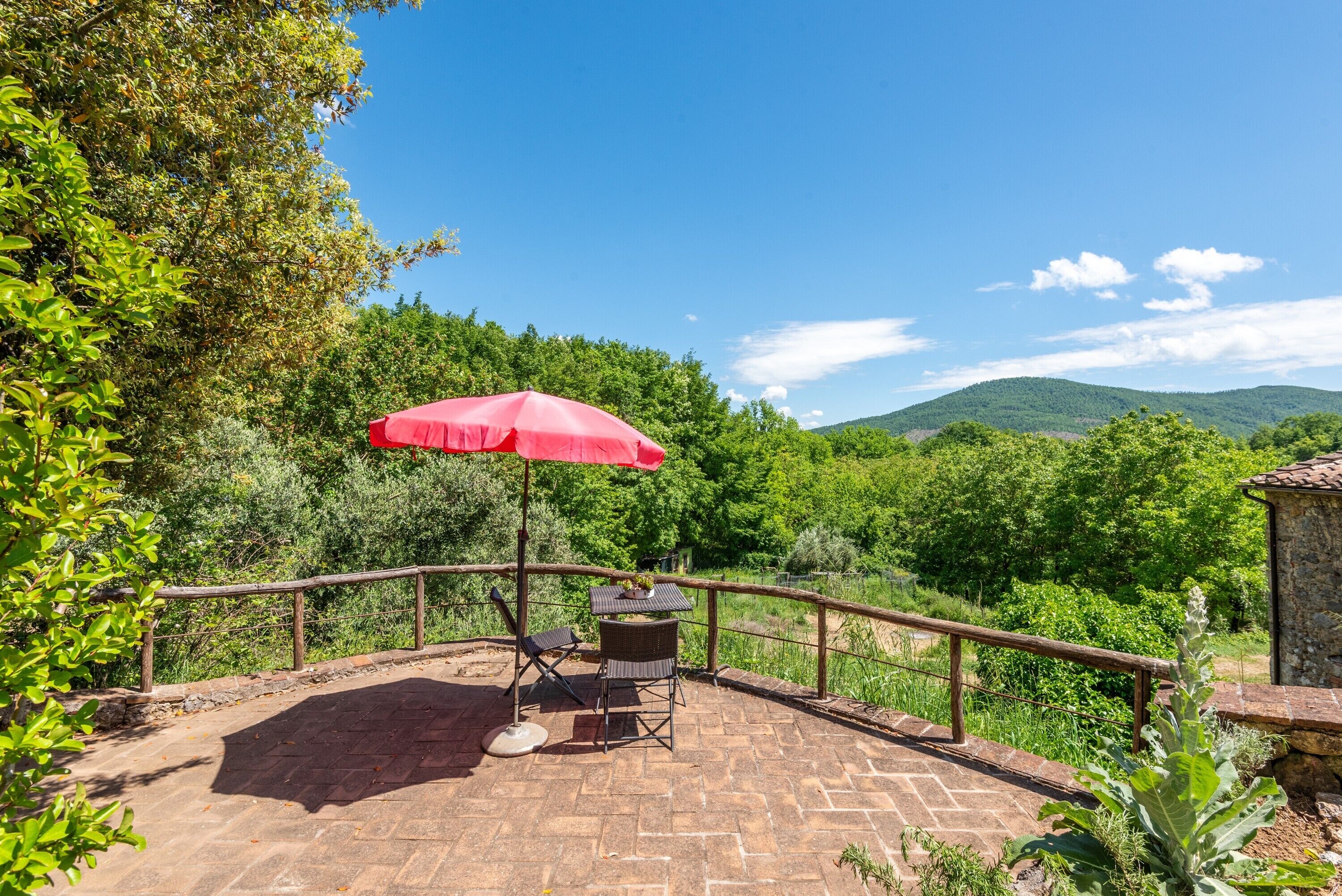 Eine Terrasse mit einem roten Sonnenschirm, zwei Stühlen und einem Tisch mit Blick auf eine üppige, grüne Landschaft und Hügel.