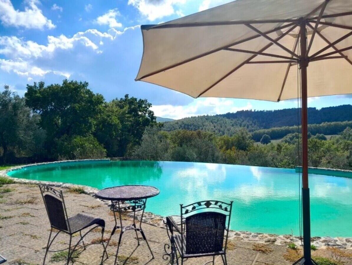 Ein klares, türkisblaues Schwimmbecken mit einer umlaufenden Steinterrasse, einem Tisch mit Stühlen und einem Sonnenschirm.