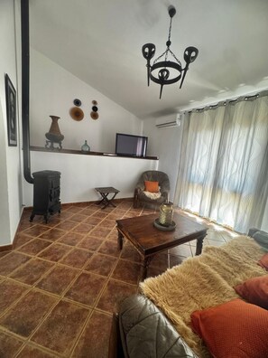 Ein Wohnzimmer mit einer Couch, einem Holztisch, einem Fernseher auf einem Regal, einem Kamin und einer Chandelier.