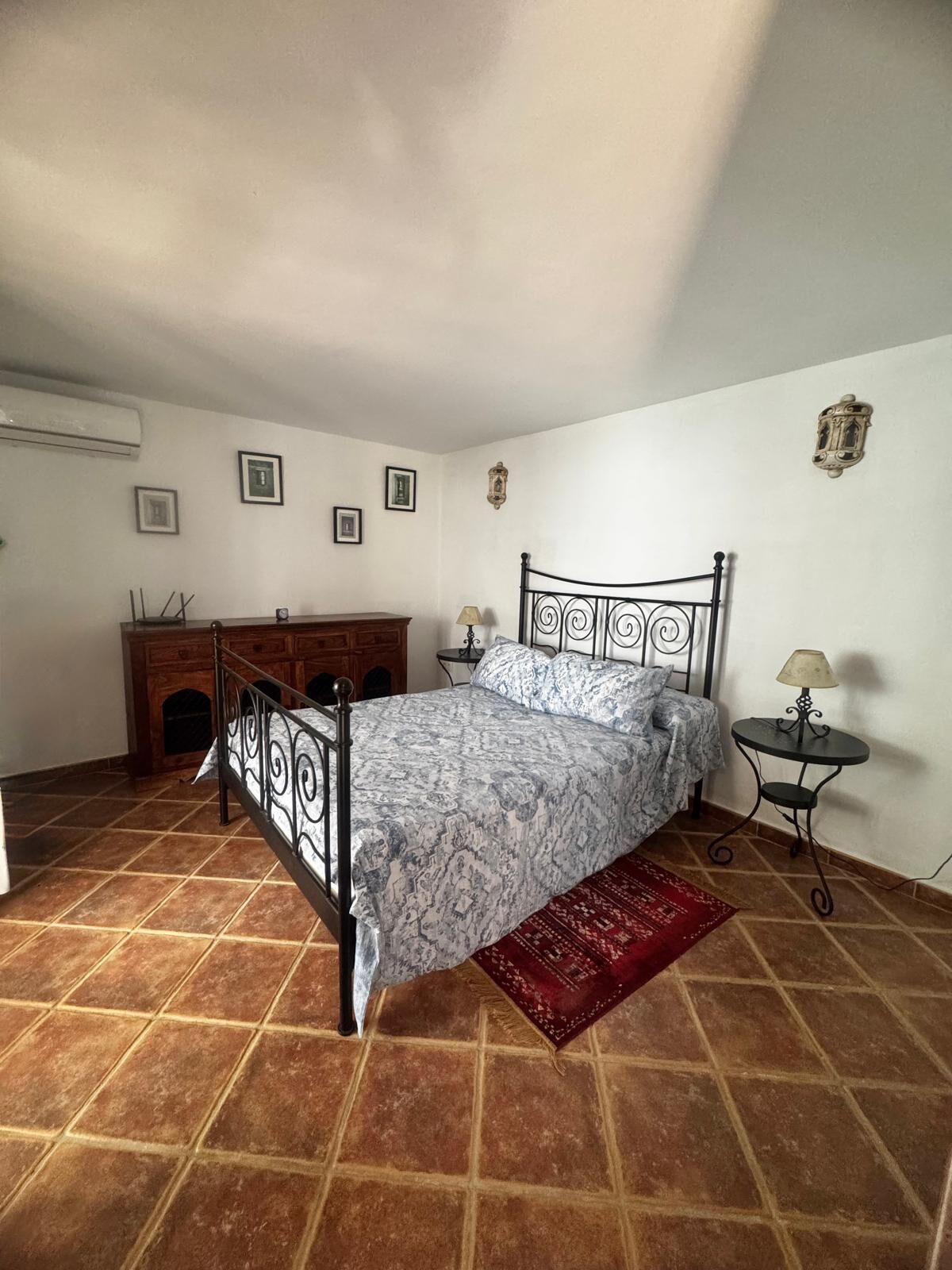 Un dormitorio con estructura de cama metálica, ropa de cama estampada, una alfombra roja, una mesita con una lámpara y una chimenea.