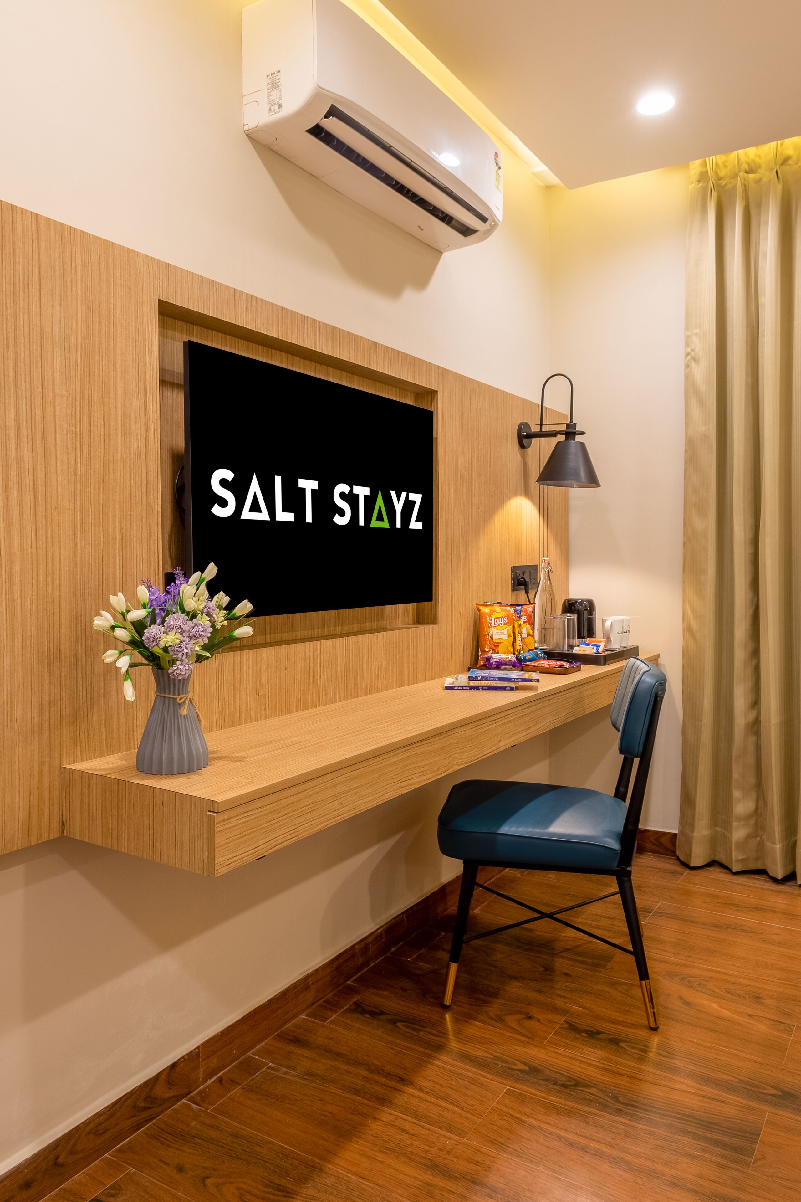 Habitación de hotel con televisión de pared, escritorio con silla, un jarrón de flores y lámpara de pared.