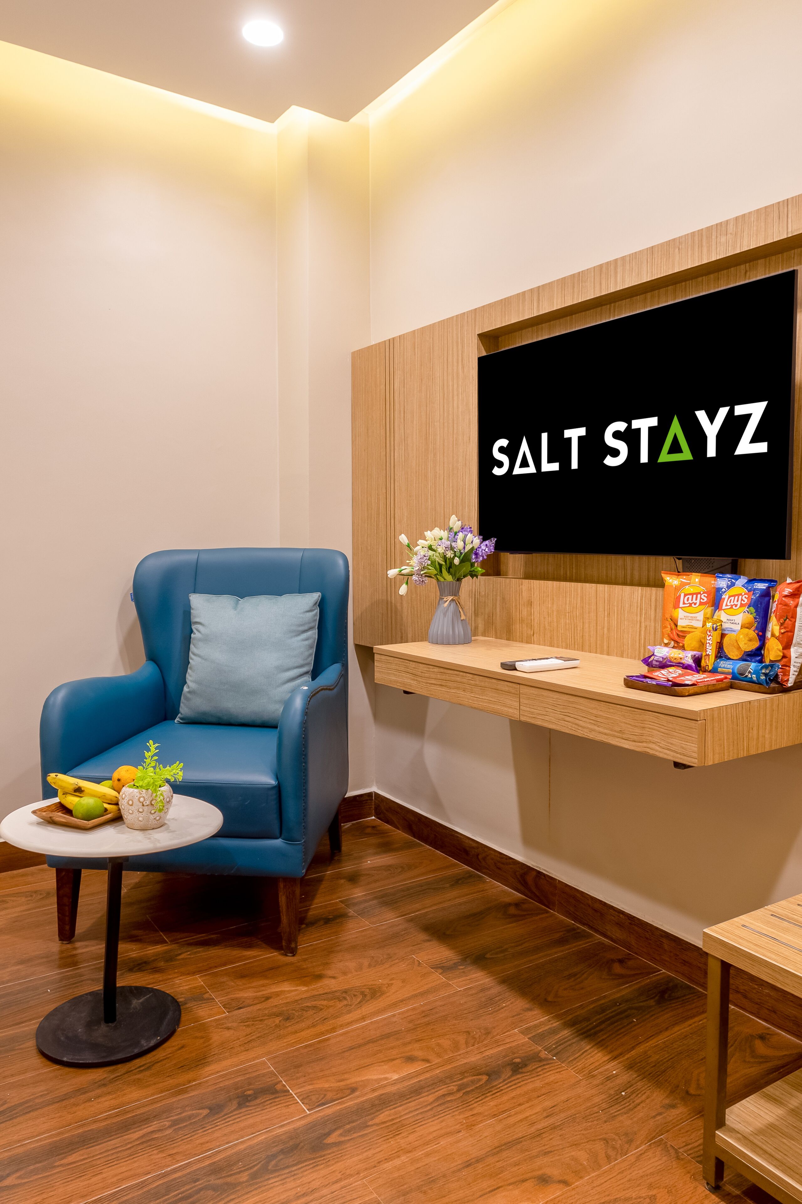 A room with a blue chair, a small table with fruits, a wall-mounted TV displaying 'SALT STAYZ', and snack bags on a shelf.
