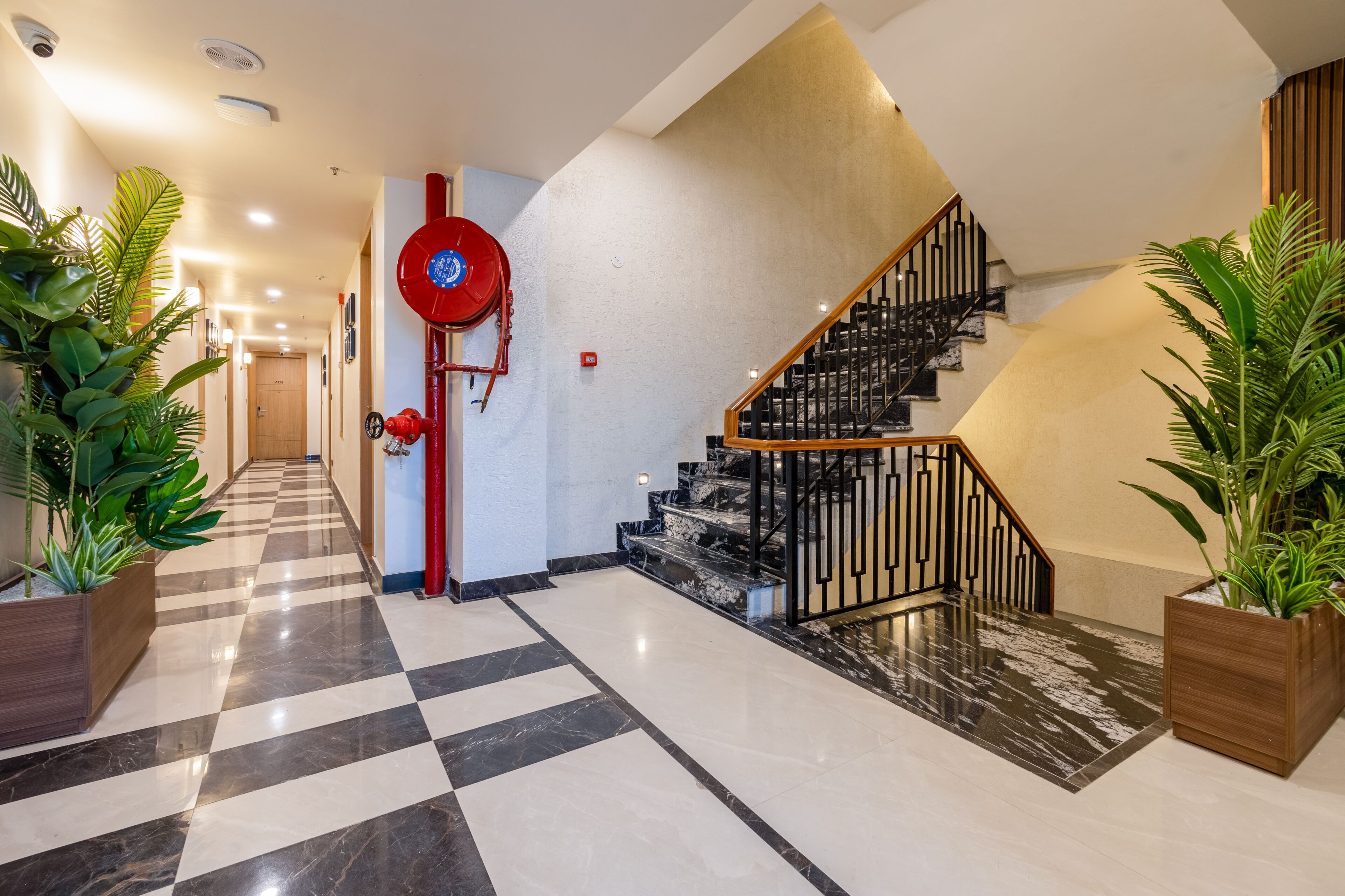 Vestíbulo de hotel con piso a cuadros, una manguera contra incendios roja, una escalera con barandas negras y plantas en macetas.