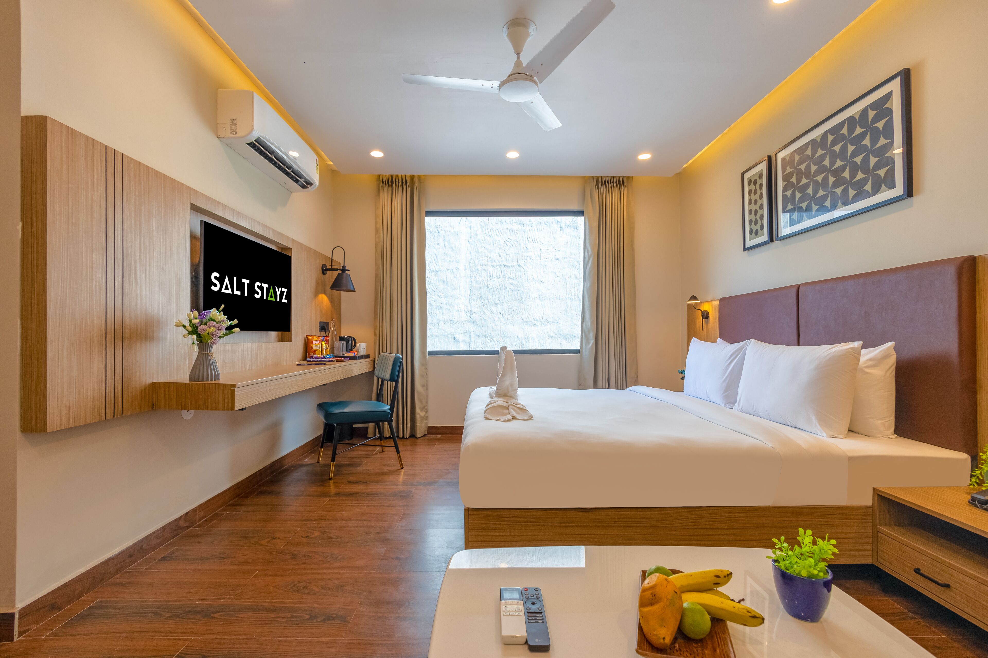 Habitación de hotel con cama, televisor montado en la pared, ventilador de techo y ventana con cortinas.