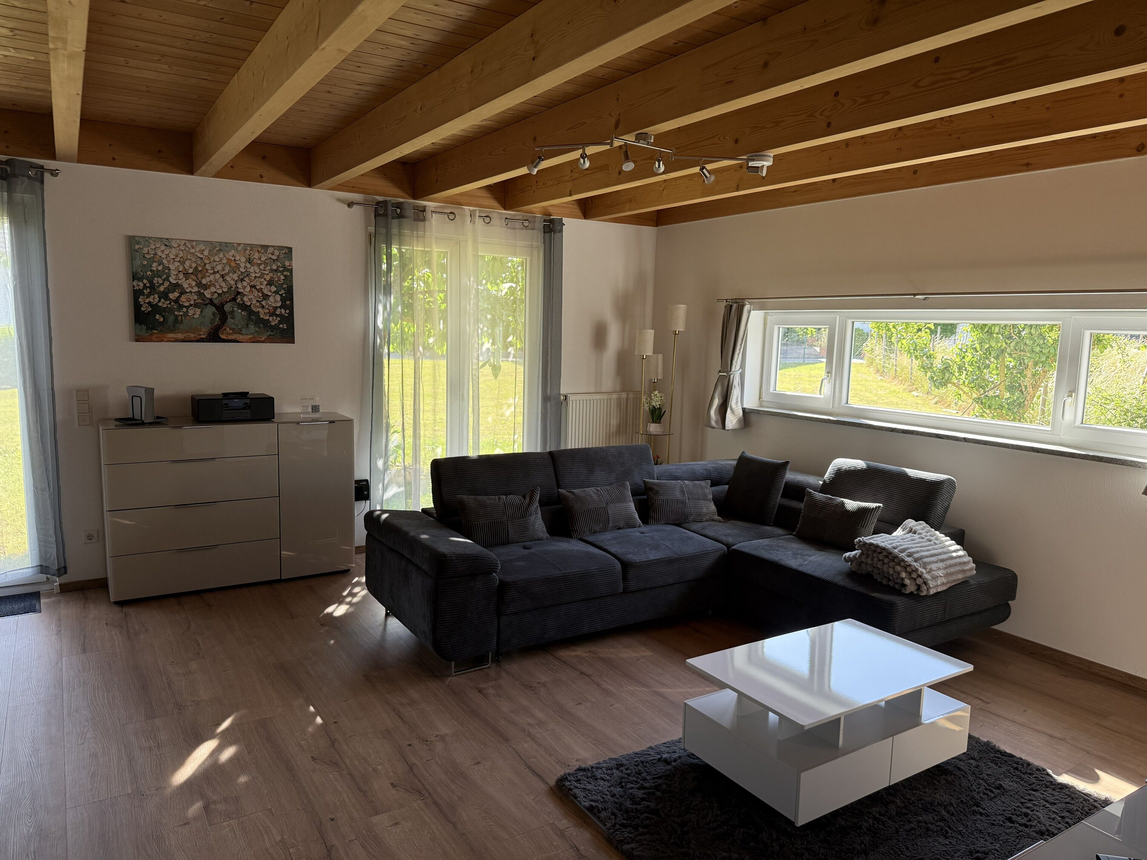 Ein modernes Wohnzimmer mit einem Ecksofa, einem weißen Couchtisch und einem großen Fenster mit Blick auf Grünflächen.