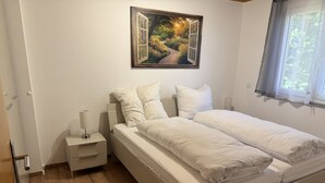 Ein Schlafzimmer mit einem Bett, Nachttischen, Vorhängen vor einem Fenster und einem Bild von einer Gartenansicht.