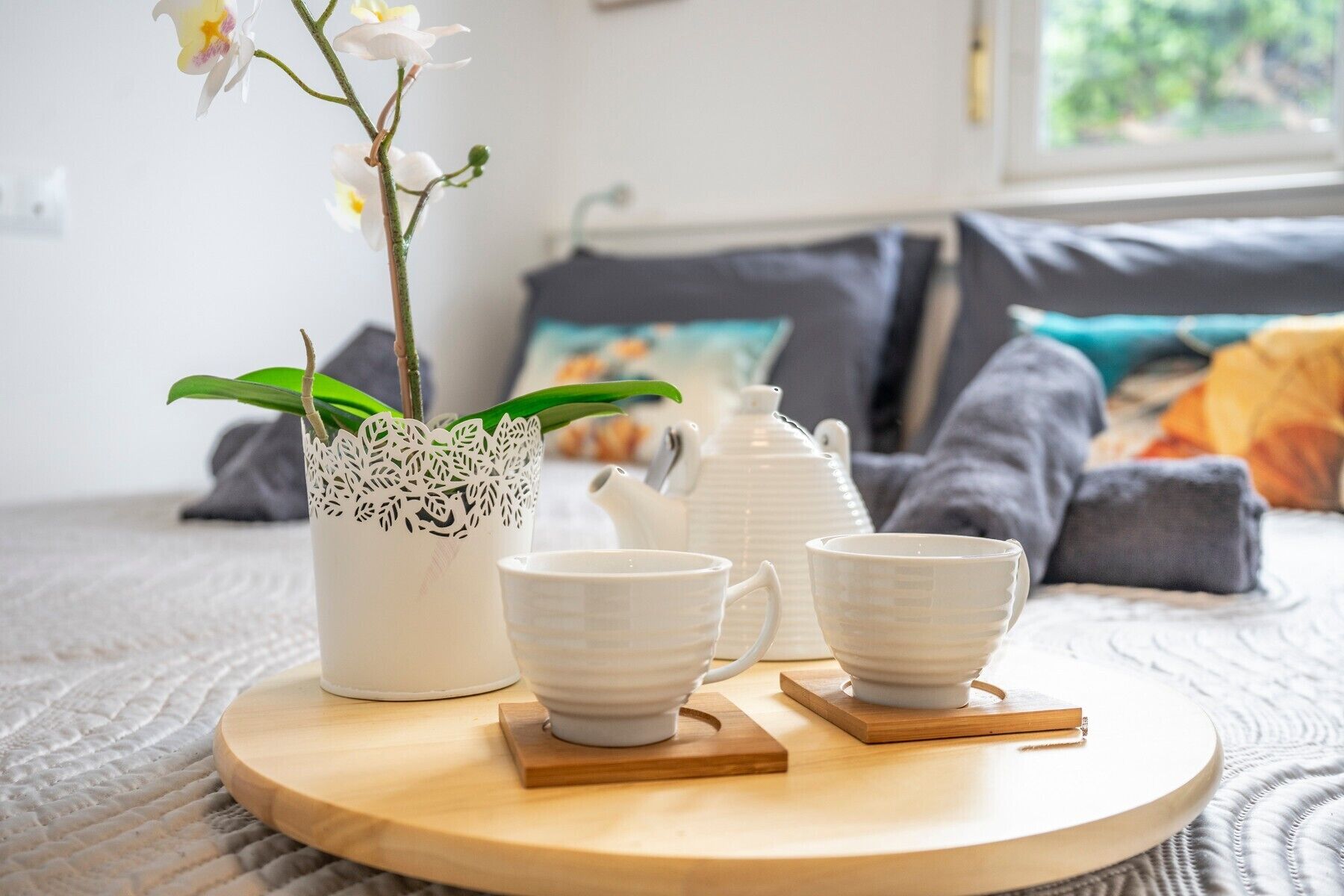 A cozy room with a bed, pillows, a white orchid plant, a white teapot, and two white cups on a wooden tray.