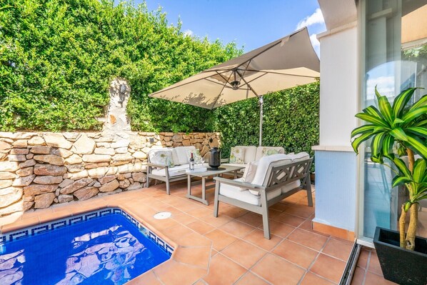 A patio with a stone wall, a pool, outdoor furniture, and a large umbrella providing shade.