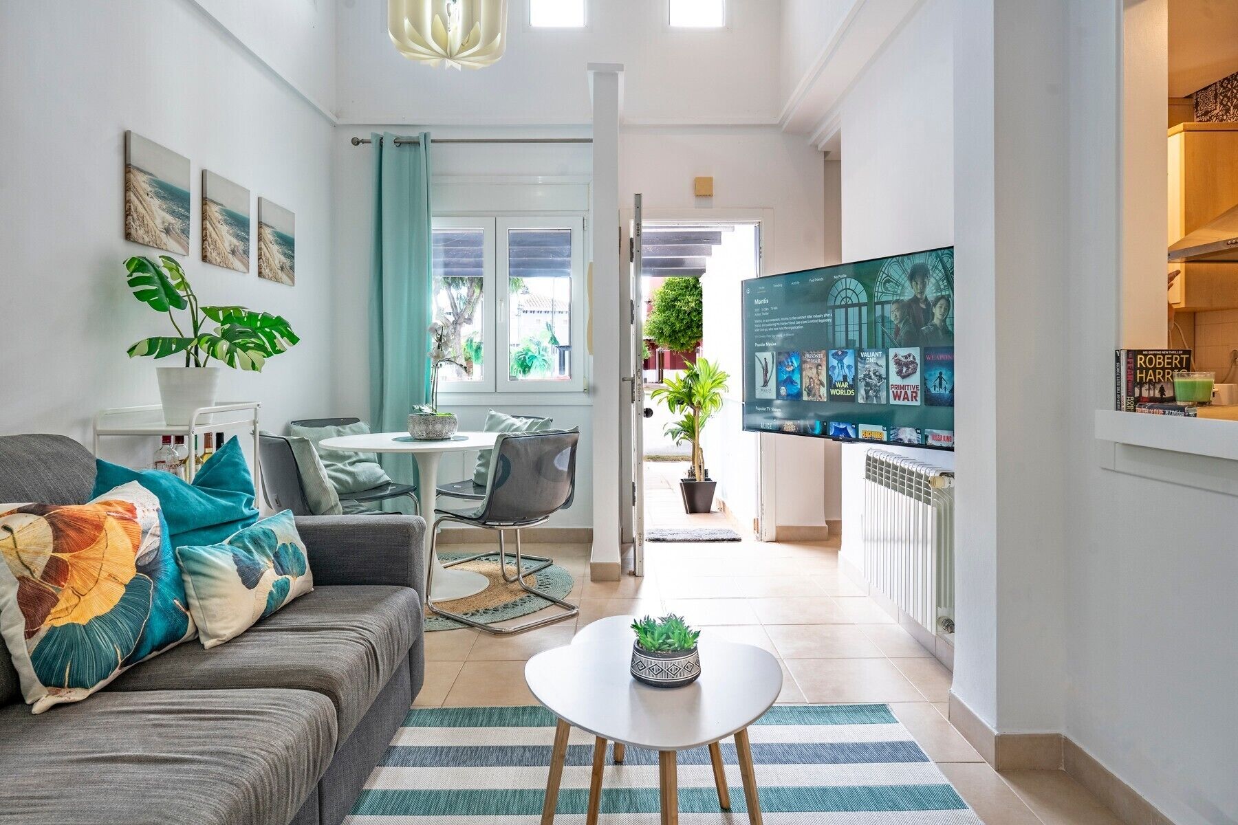 A modern living room with a sofa, coffee table, and TV. There is a dining area with chairs and a table. The room has a large window and a view of the outside.