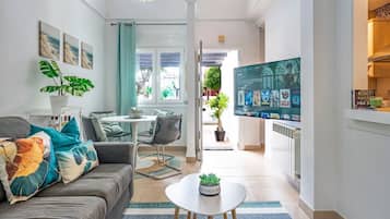 A modern living room with a sofa, coffee table, and TV. There is a dining area with chairs and a table. The room has a large window and a view of the outside.