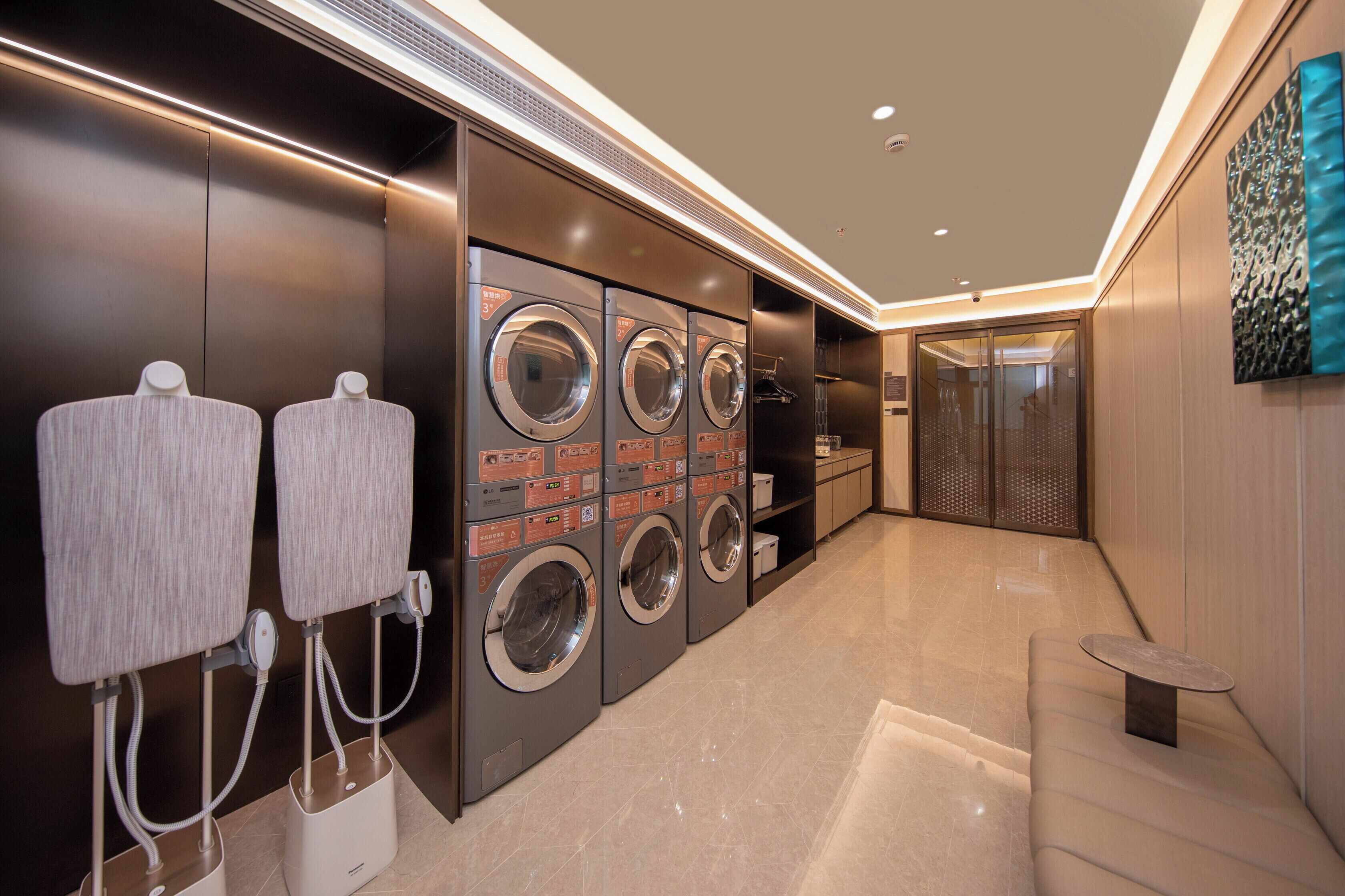 A modern laundry facility with multiple washing machines and dryers, a seating area, and a glass door leading to another room.