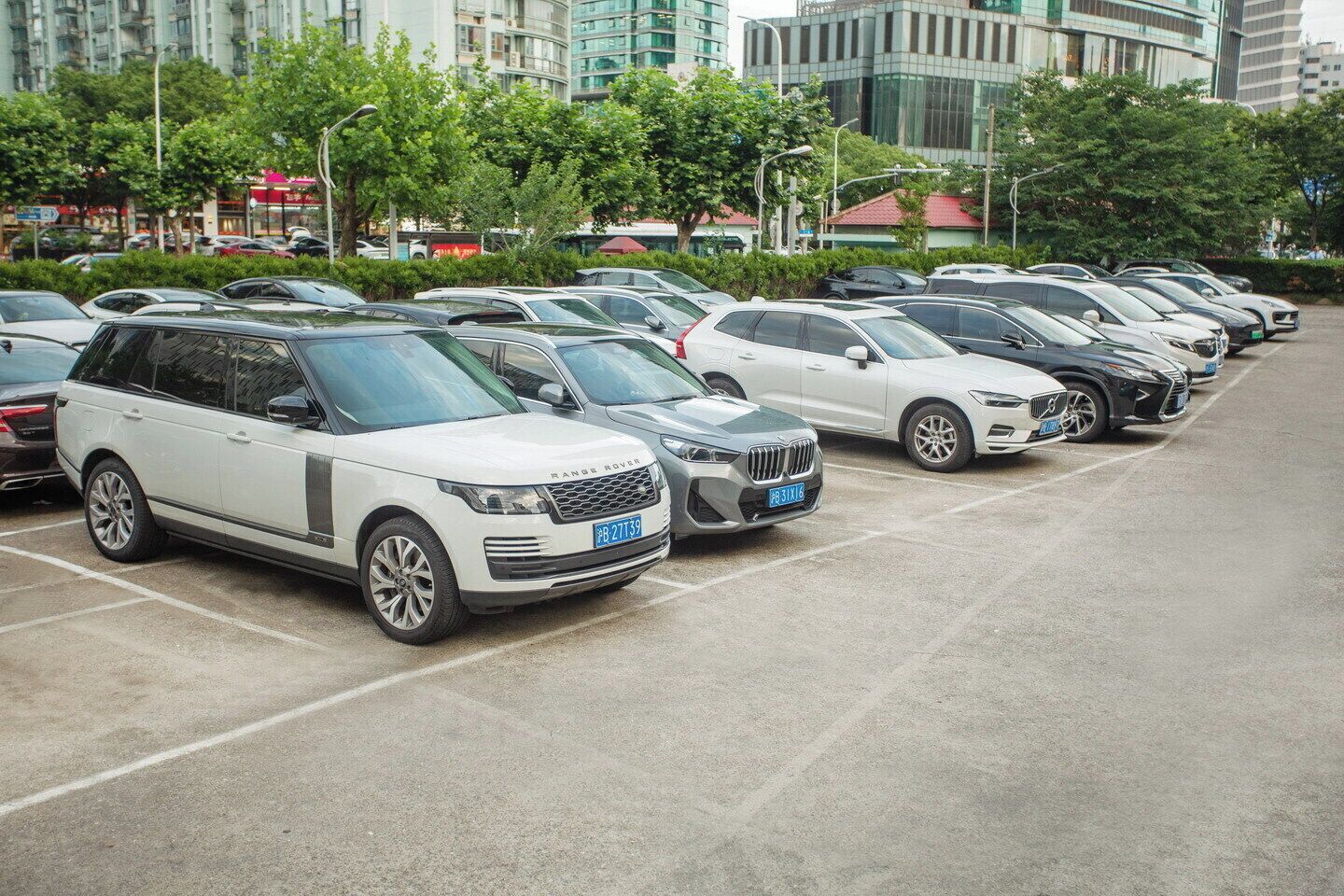 A parking lot with multiple cars, including a white Range Rover and a silver BMW, with buildings and trees in the background.