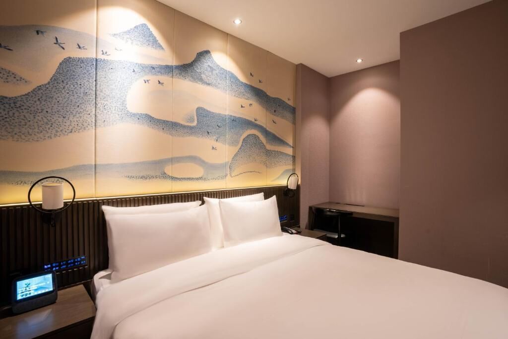 Quarto de hotel com cama, mesa de cabeceira com abajur, obra de arte na parede e painel de controle.