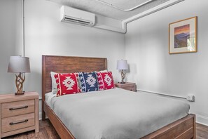 Un dormitorio con cabecera de madera, cama con almohadones estampados, lámparas de noche, aire acondicionado instalado en la pared y un cuadro enmarco.