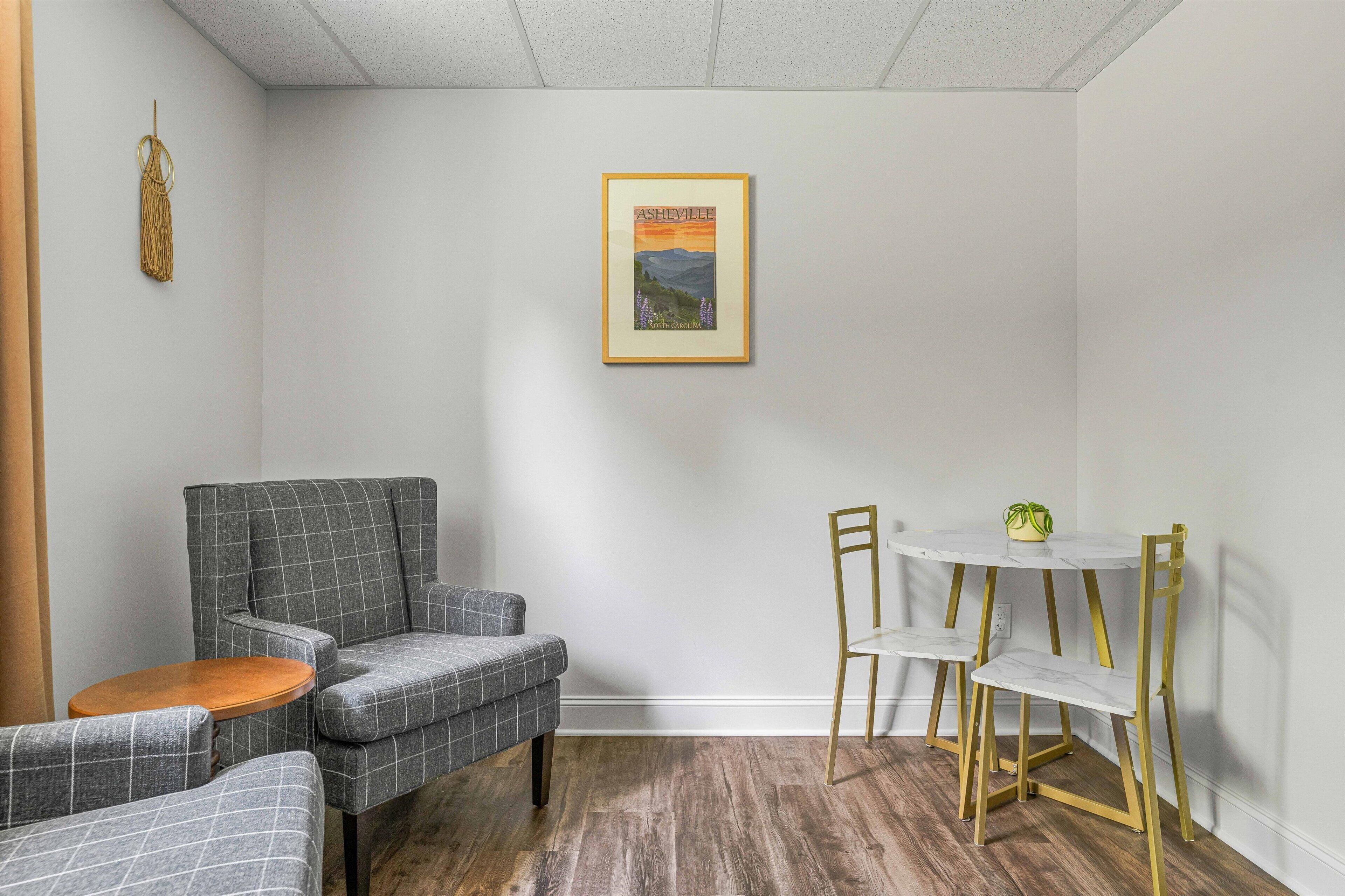 Una habitación pequeña con un sillón tapizado gris, una mesa redonda de madera, una mesa redonda blanca con una silla dorada y un cuadro en la pared.