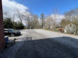 A gravel parking area with a black car parked on the left, a red car in the background, and a building with a brick facade on the left. - Sabel #004 | Downtown Asheville twin suite with bath, Wi-Fi, AC, and parking. (Asheville)