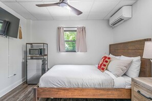 A bedroom with a bed, a ceiling fan, an air conditioner, a window with curtains, a TV, and a kitchenette with a microwave. - Sabel #005 | Downtown Asheville with queen bed (Asheville)