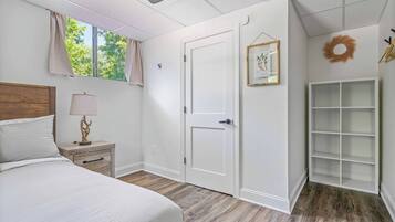 A bedroom with a bed, bedside table, lamp, window with curtains, door, and a built-in shelving unit.