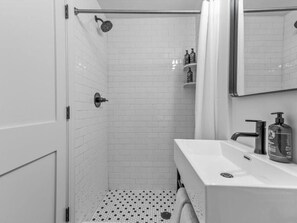 A bathroom with white tiled walls, a shower with a handheld showerhead, a sink with a soap dispenser, and a towel hanging on the wall. - Sabel #103 Downtown Asheville king suite with Wi-Fi, parking, and kitchenette (Asheville)