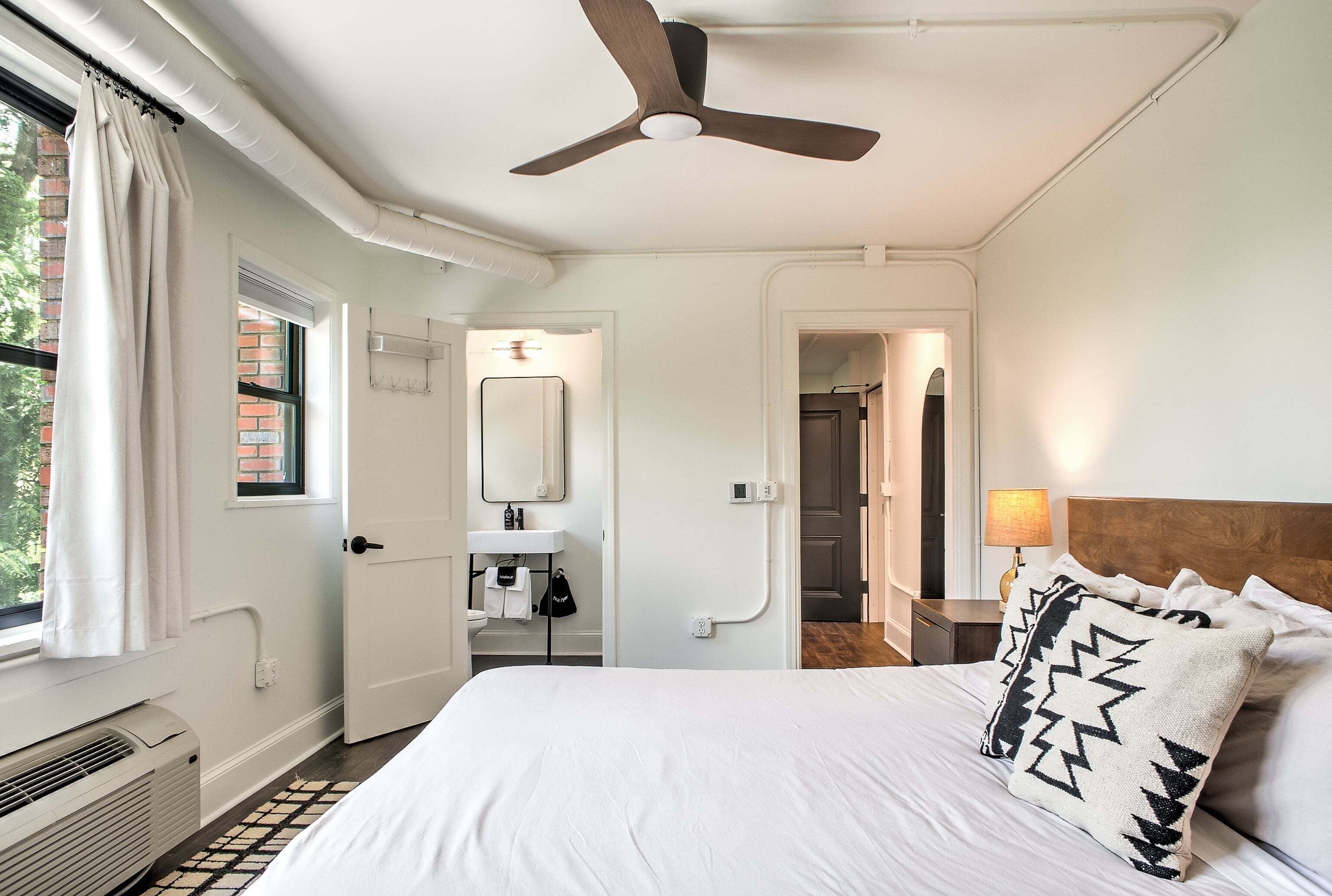 A hotel room with a bed, a ceiling fan, a window with curtains, a bathroom with a mirror and sink, and a hallway leading to another room.