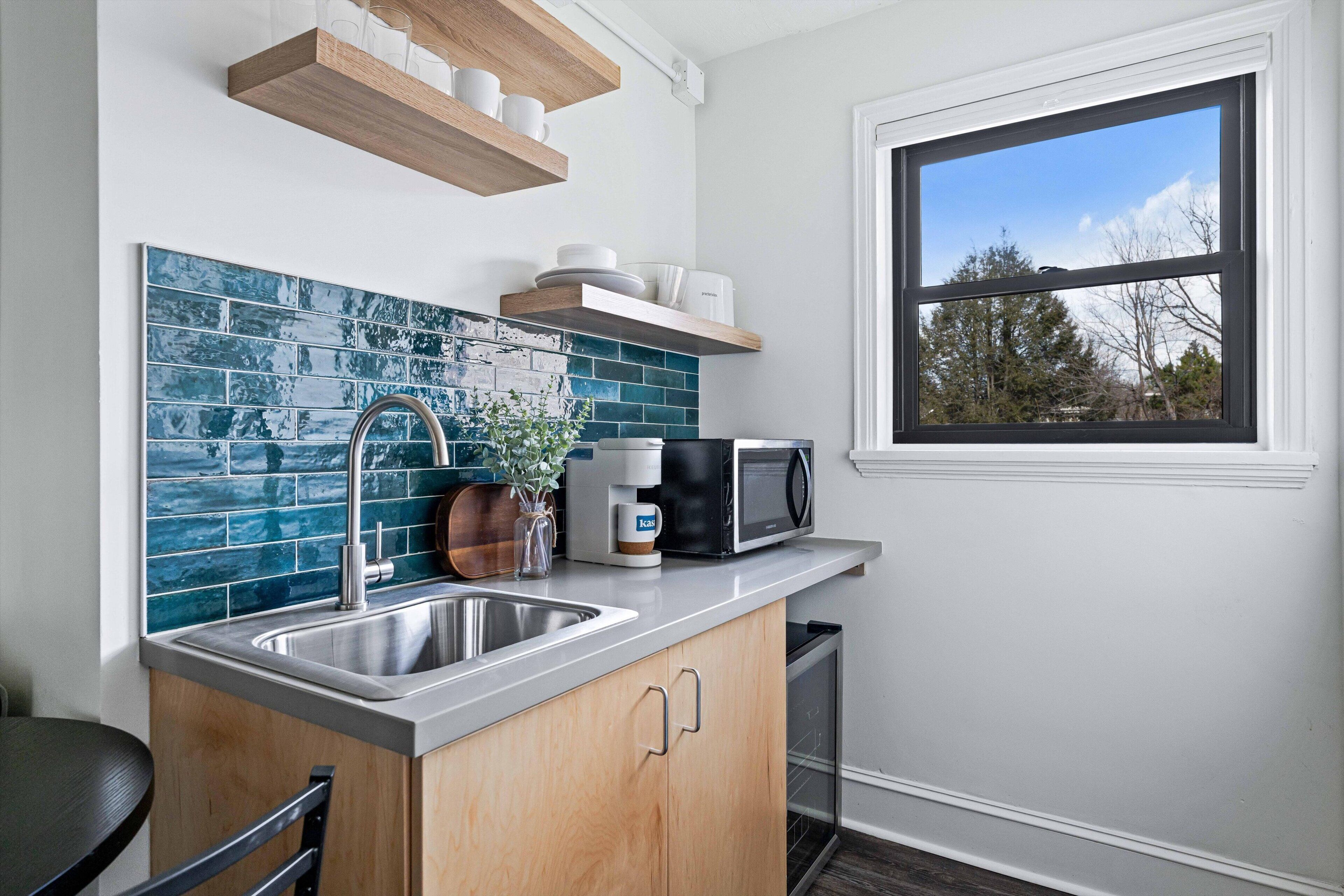 A kitchen with a blue tile backsplash, a stainless steel sink, wooden cabinets, a microwave, and a window with a view of trees.