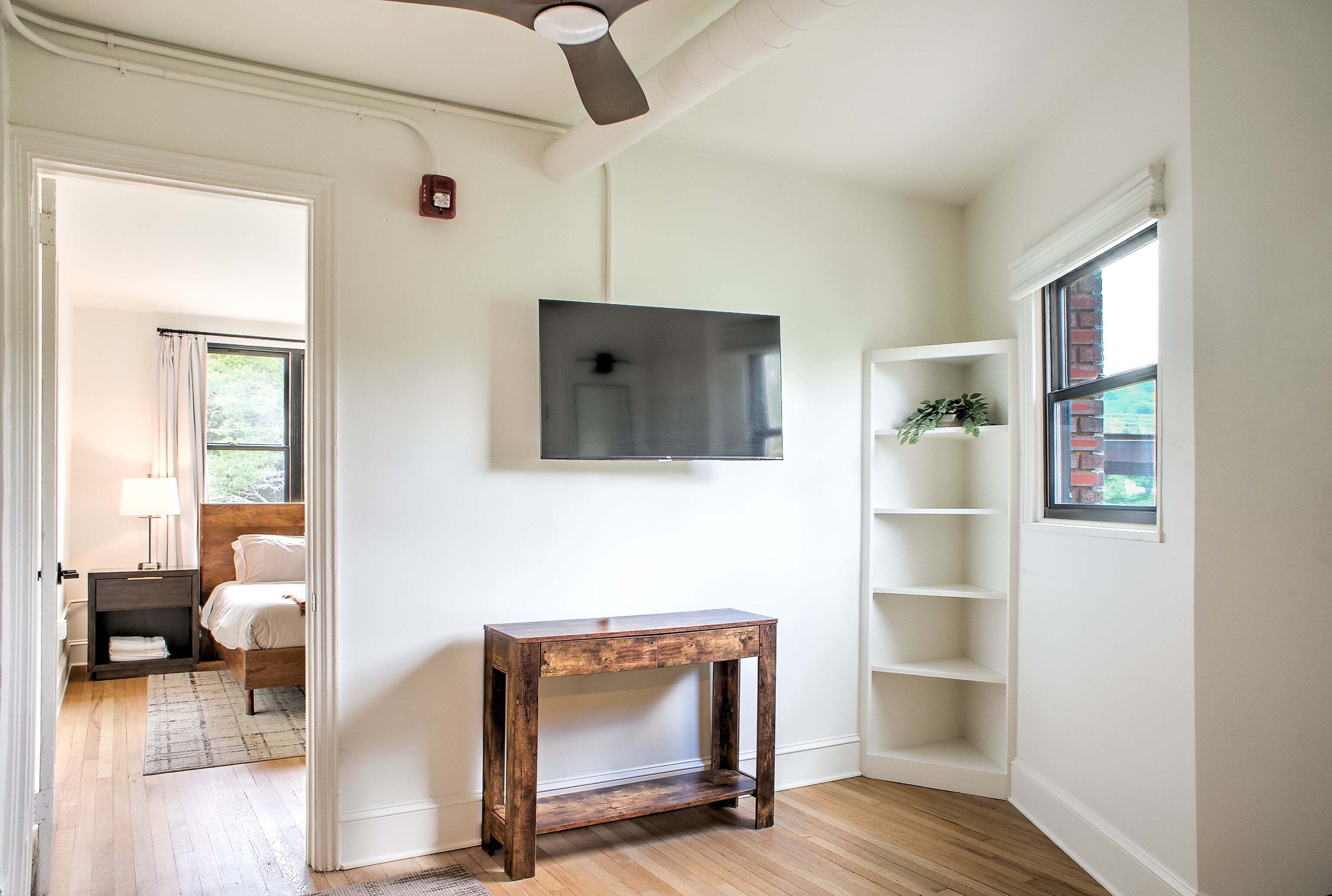 A room with a bed, a wooden table, a TV mounted on the wall, and a window with a view of greenery.