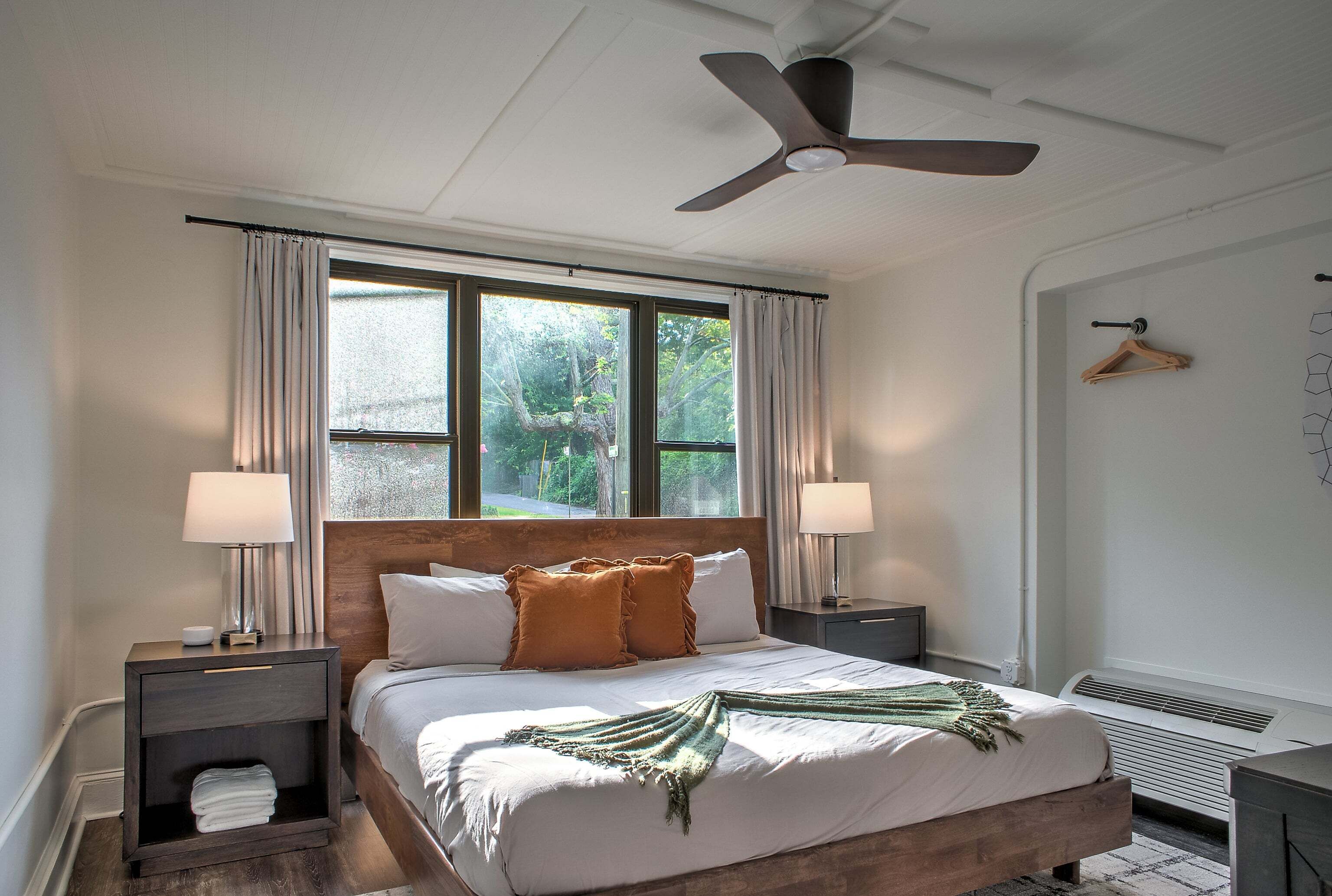 A bedroom with a bed, two bedside tables, a ceiling fan, a window with curtains, and a wall-mounted heater.