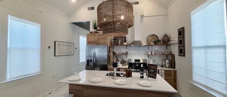 A modern kitchen with a central island, bar stools, a hanging light fixture, and a 'BE OUR GUEST' sign.
