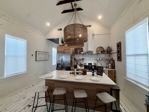 Een moderne keuken met een centraal eiland, barstoelen, een hanglamp en een bord met de tekst 'BE OUR GUEST'.